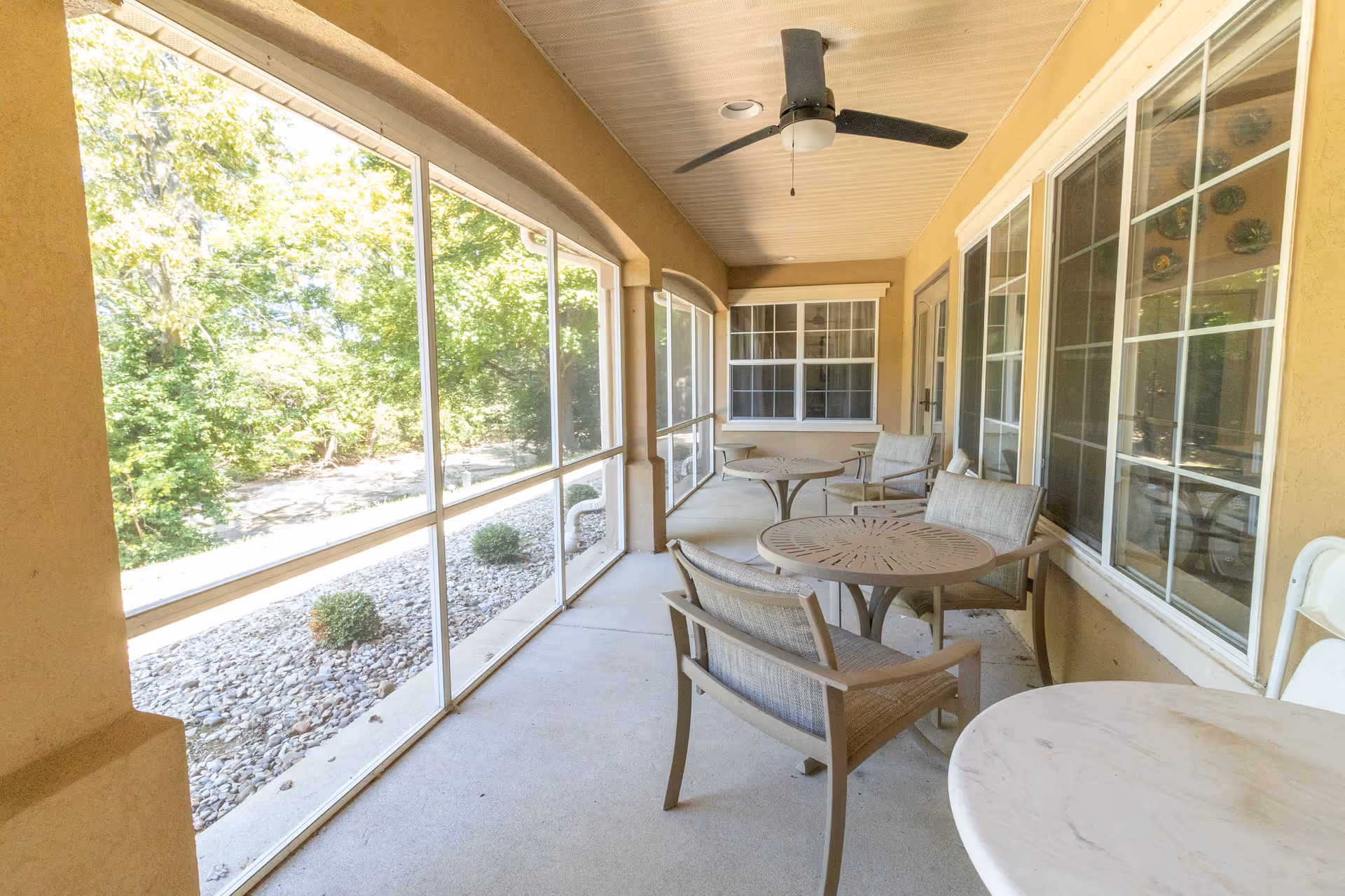 A screened-in porch area with several round tables and cushioned chairs arranged along the wall with large windows. A ceiling fan is mounted on the porch ceiling. Outside the screened area, there is a garden bed with rocks and small bushes, and trees are visible in the background.