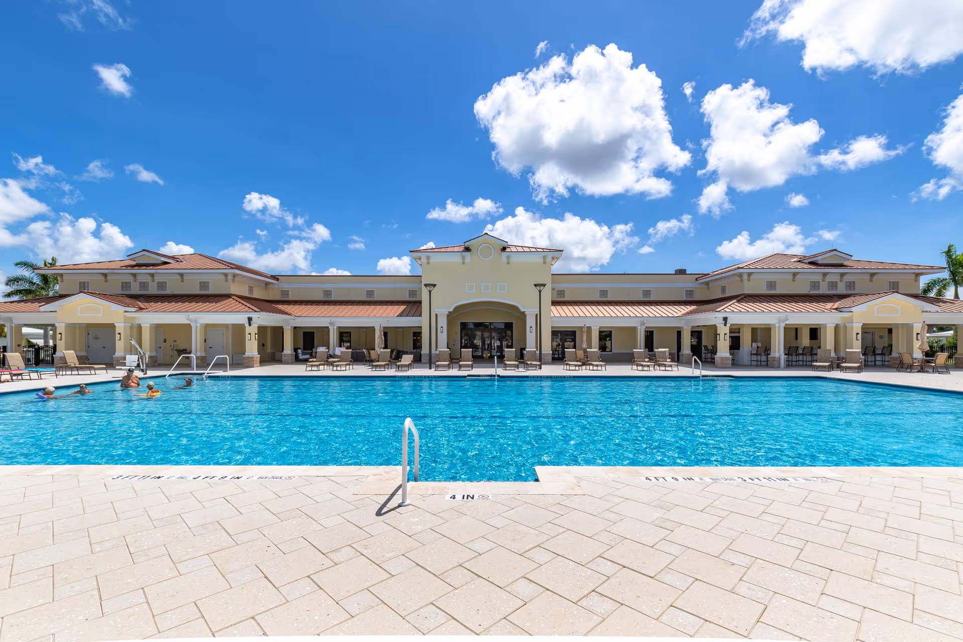 Outdoor swimming pool with several people swimming and lounging around it, in front of a large, light yellow building with a red-tiled roof under a bright blue sky with scattered white clouds.
