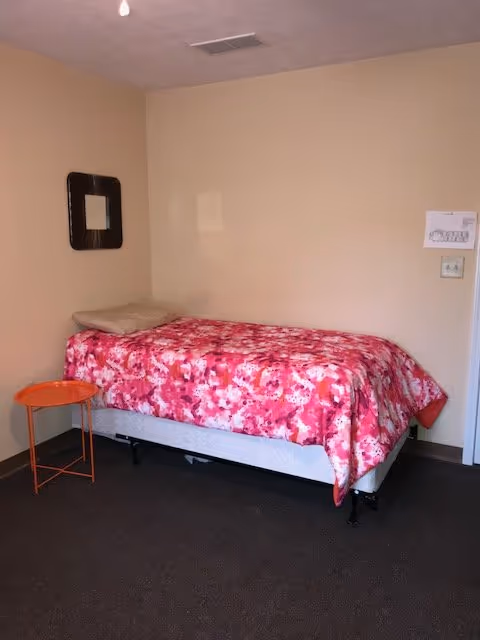 Simple single bedroom with a bed covered in a pink floral blanket, a small round orange side table, and a wall mirror.
