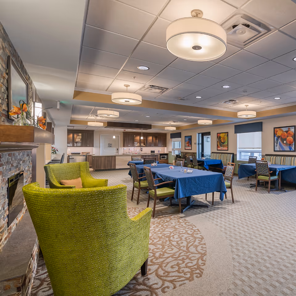 Spacious communal dining room with tables covered in blue tablecloths, green armchairs by a stone fireplace, and modern ceiling lights.