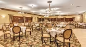 A spacious dining room with multiple round tables covered with white tablecloths and set with napkins and silverware. The room features patterned carpet flooring, upholstered chairs with wooden frames, chandeliers hanging from the ceiling, and windows with curtains along the walls.