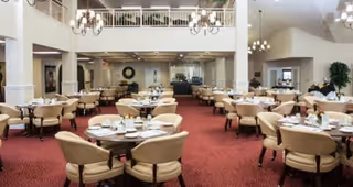 Spacious dining room with multiple round tables set with plates, glasses, and napkins. The room features beige upholstered chairs, red carpet flooring, chandeliers hanging from the ceiling, and a balcony railing overlooking the area.
