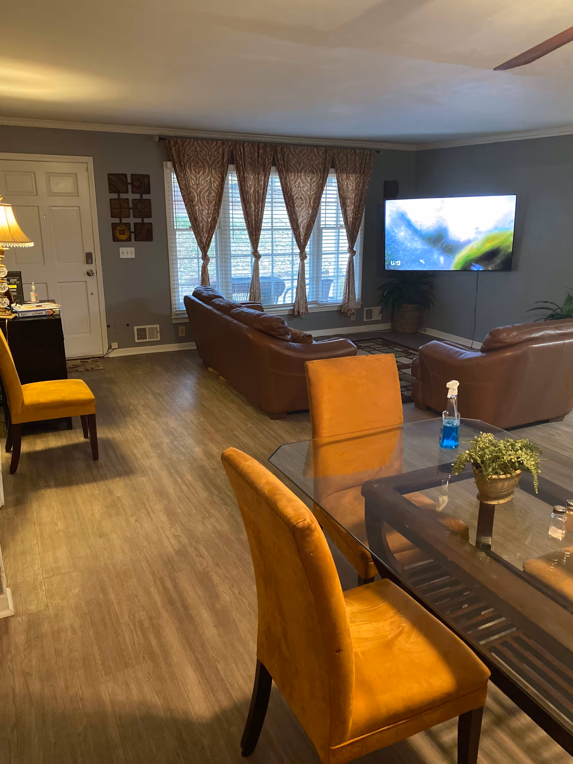A living room area with brown leather sofas facing a wall-mounted flat screen TV. The room has large windows with patterned curtains, a glass dining table with mustard yellow chairs, a wooden floor, and a lamp on a side table near the entrance door.