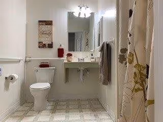 A clean and well-lit bathroom featuring a toilet, a wall-mounted sink with a mirror above it, a towel hanging on the wall, a shower curtain with a floral pattern, and a decorative wall hanging.
