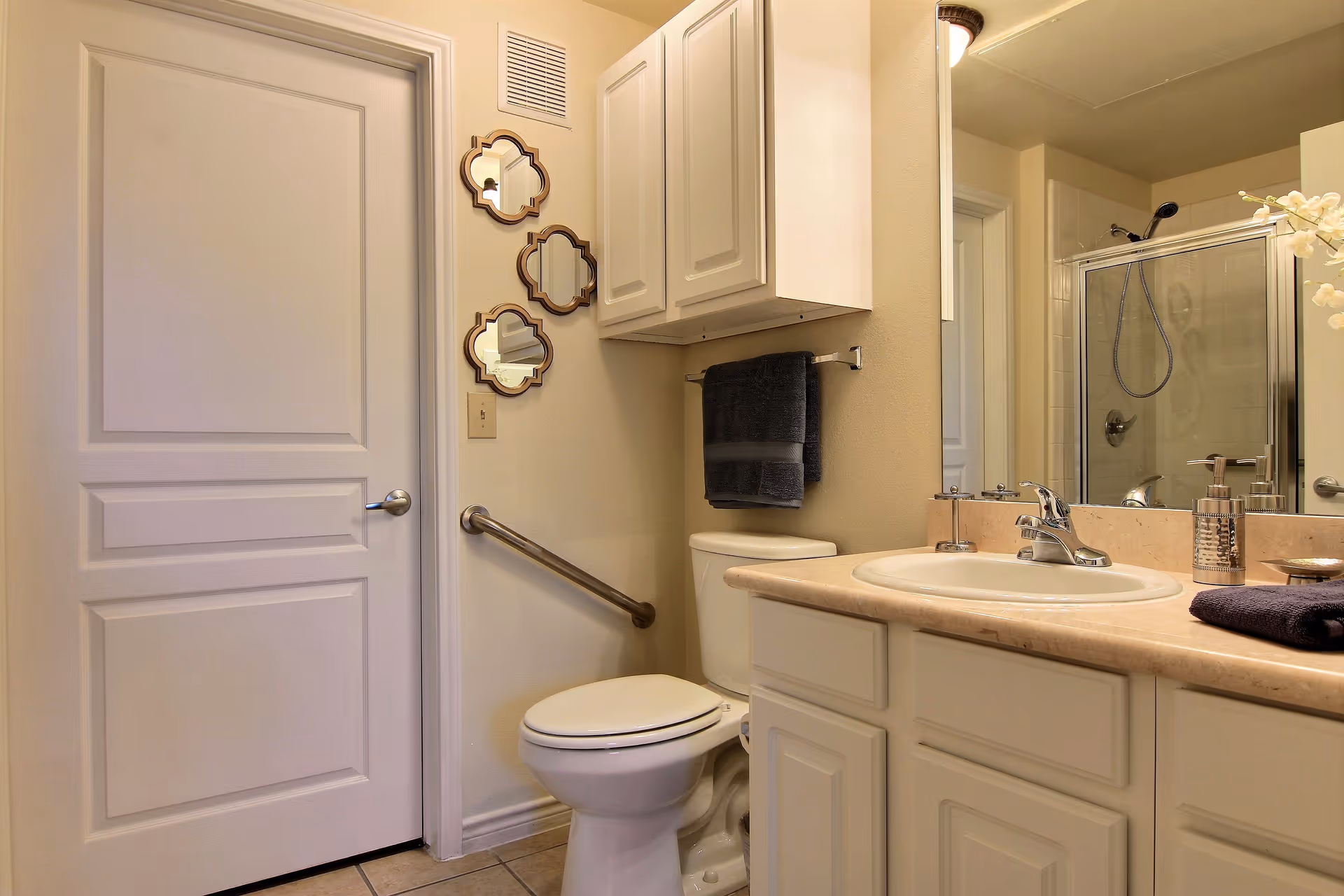 A clean bathroom featuring a white toilet with a grab bar on the wall beside it, a beige countertop with a sink, a large mirror above the sink, white cabinets both above and below the countertop, a towel rack with dark towels, and a glass-enclosed shower in the background. Three decorative mirrors are mounted on the wall next to a closed white door.