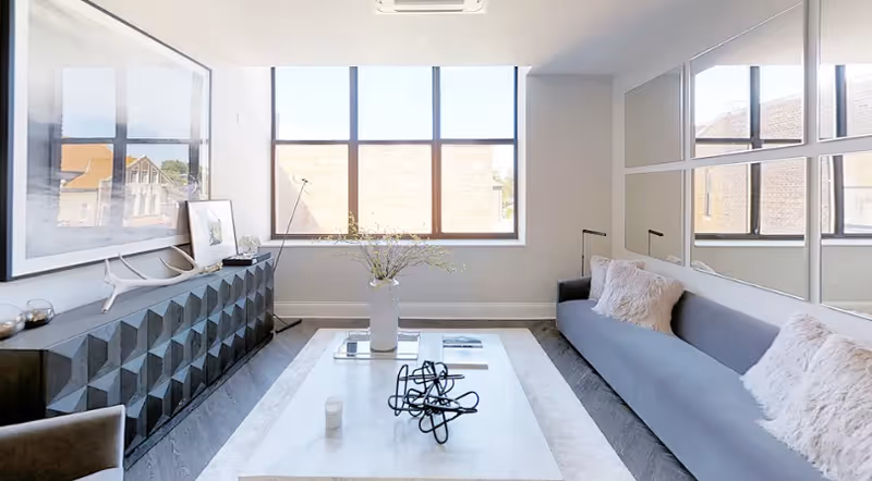 A modern living room with a large window letting in natural light. The room features a long gray sofa with two fluffy pillows, a white rectangular coffee table with decorative items including a vase with flowers and a black abstract sculpture. On the left side, there is a black geometric-patterned cabinet with framed artwork and decorative objects on top. The wall behind the sofa has a grid of square mirrors.