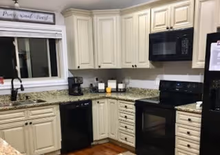 A kitchen with cream-colored cabinets, granite countertops, a black dishwasher, black stove with oven, black microwave, black refrigerator, a coffee maker, and various kitchen items on the counter. There is a window above the sink with a sign above it that reads 'My sweet home'.