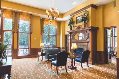 Sunlit communal lounge with upholstered chairs and sofas arranged around a decorative fireplace beneath tall windows with yellow draped curtains.