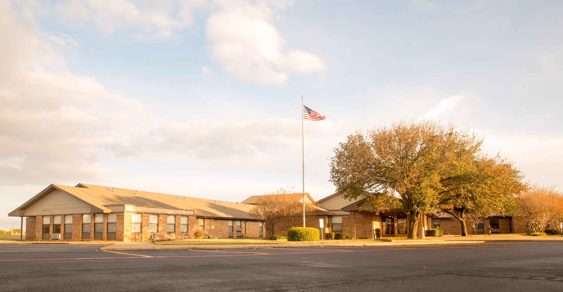 Exterior view of a single-story brick building with a pitched roof, surrounded by trees with autumn foliage. An American flag on a flagpole stands in front of the building, and there is a large paved parking area in the foreground under a partly cloudy sky.