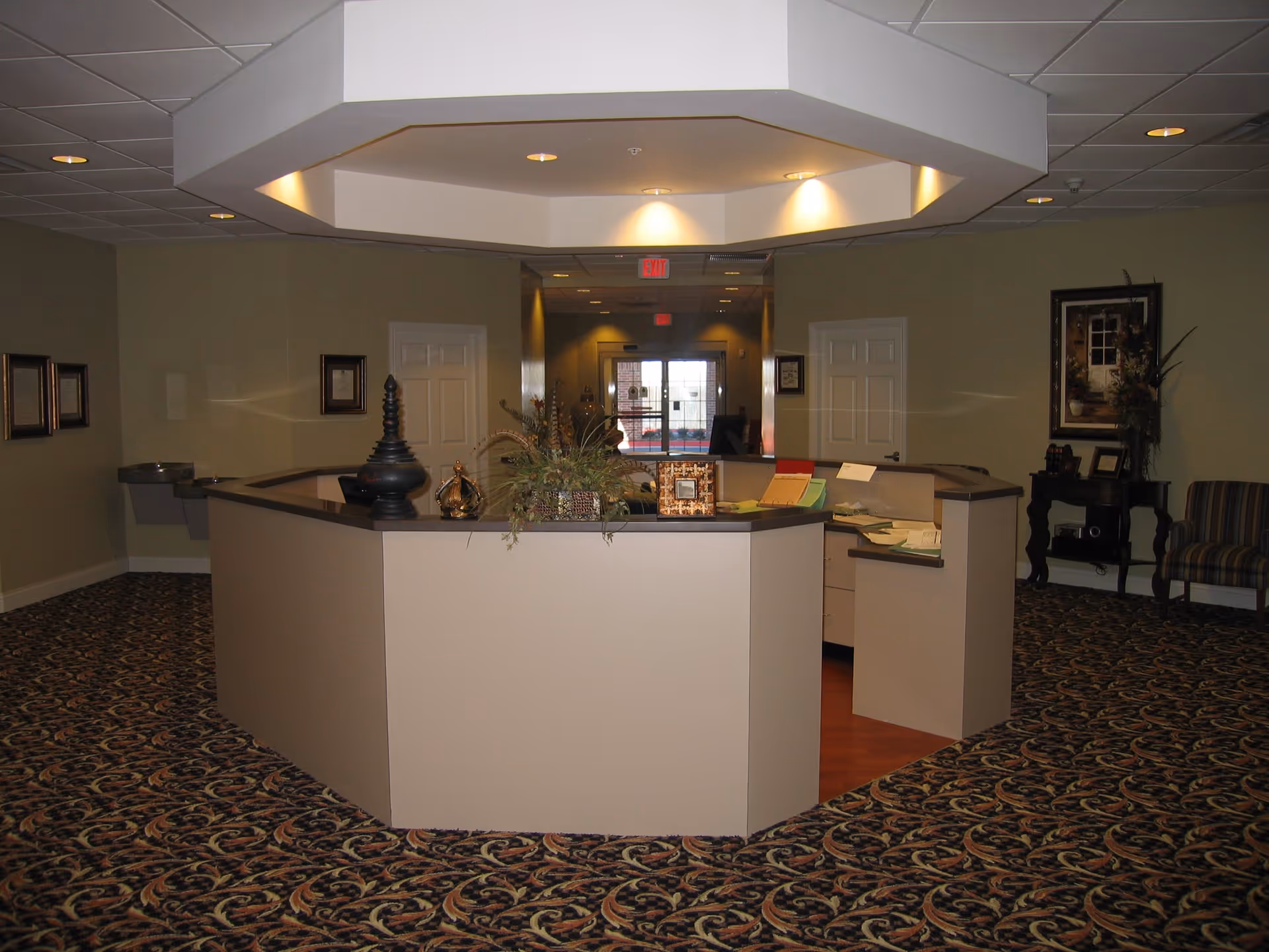 Reception area with a large octagonal desk in the center, decorated with plants and ornamental objects. The room has patterned carpet flooring, beige walls, framed pictures, and a water fountain on the left wall. There are two white doors behind the desk and a glass exit door in the background.