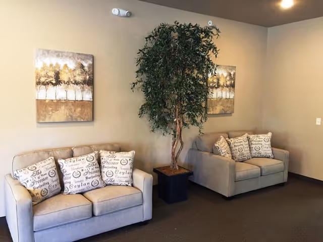 A cozy interior seating area with two beige loveseats, each adorned with three decorative pillows featuring script text. Between the loveseats is a tall potted artificial tree. Two framed paintings of trees hang on the beige wall behind the loveseats.