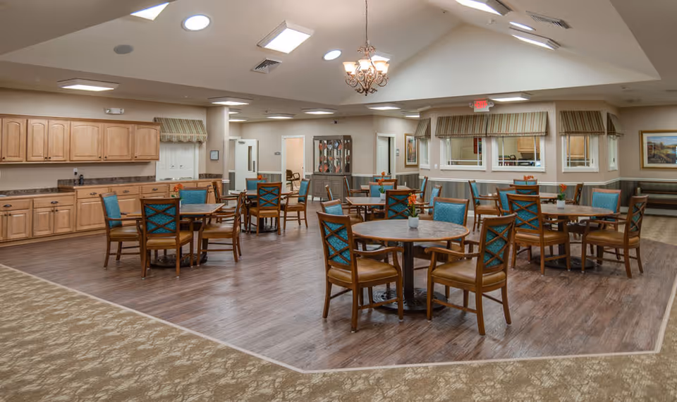 Spacious senior dining room with round wooden tables and chairs, cabinetry along one wall, and skylights in the ceiling.