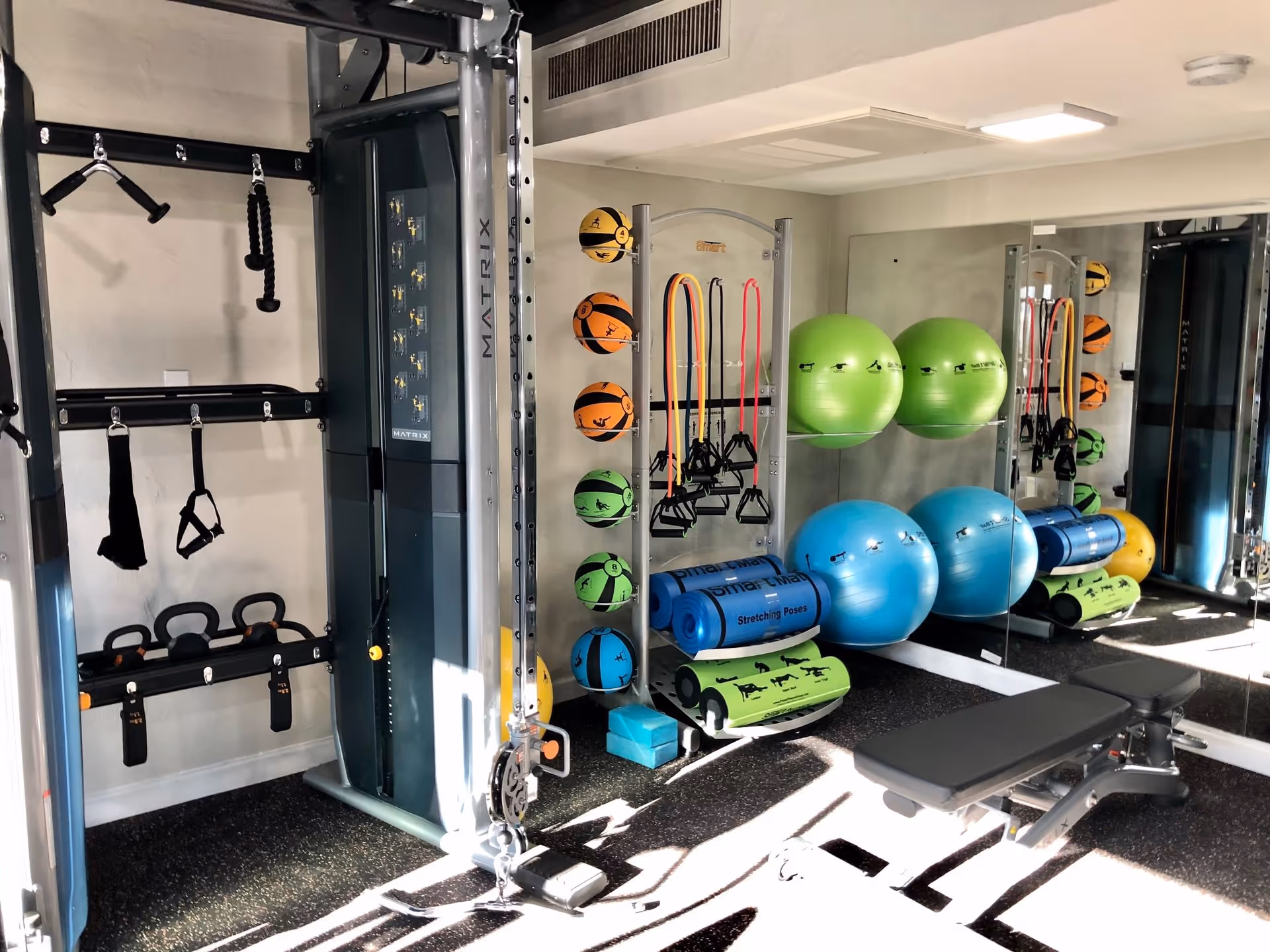 A well-lit fitness room with various exercise equipment including a multi-functional gym machine, kettlebells, resistance bands, medicine balls, yoga mats, foam rollers, and large exercise balls. There is a bench in the foreground and a large mirror on the wall reflecting the equipment.
