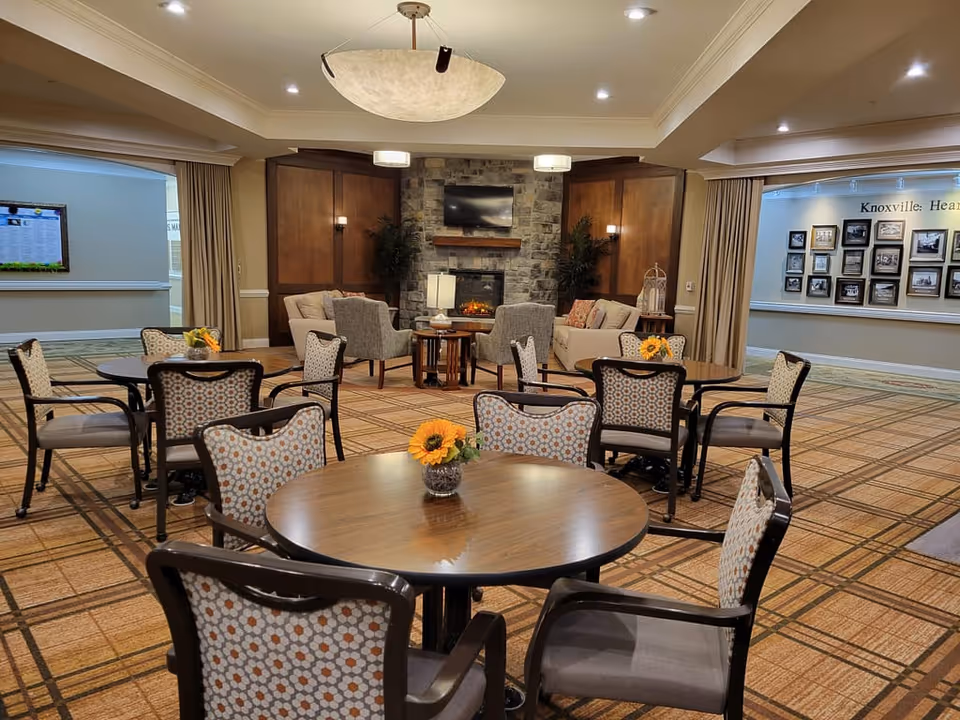 A cozy common area with round tables and patterned chairs arranged on a checkered carpet. In the background, there is a stone fireplace with a mounted TV above it, flanked by two beige sofas and armchairs. The walls have wood paneling and there are plants on either side of the fireplace. The ceiling has recessed lighting and a large central light fixture. On the right wall, there are framed pictures and a sign that reads 'Knoxville: Hea...'.