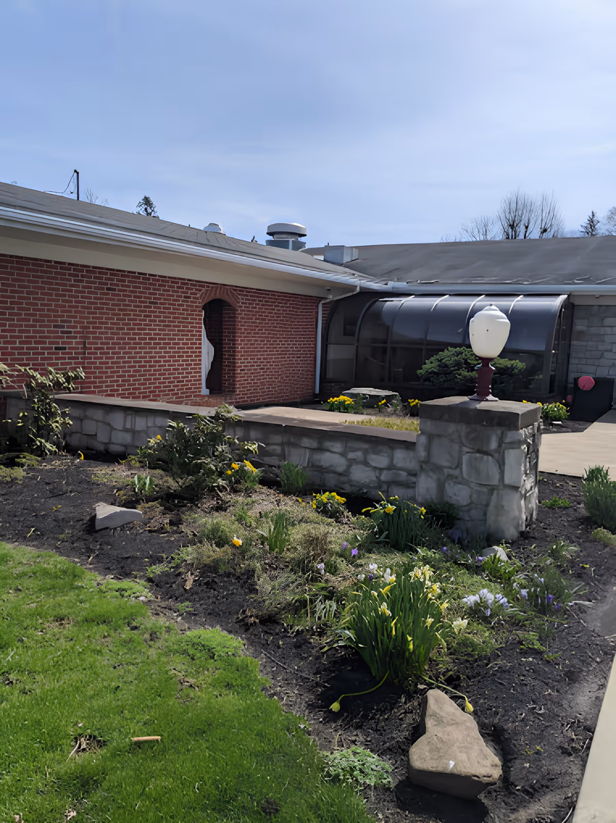 Outdoor garden area with flower beds and green grass in front of a brick and stone building with a curved glass structure and a lamp post on a stone pillar.