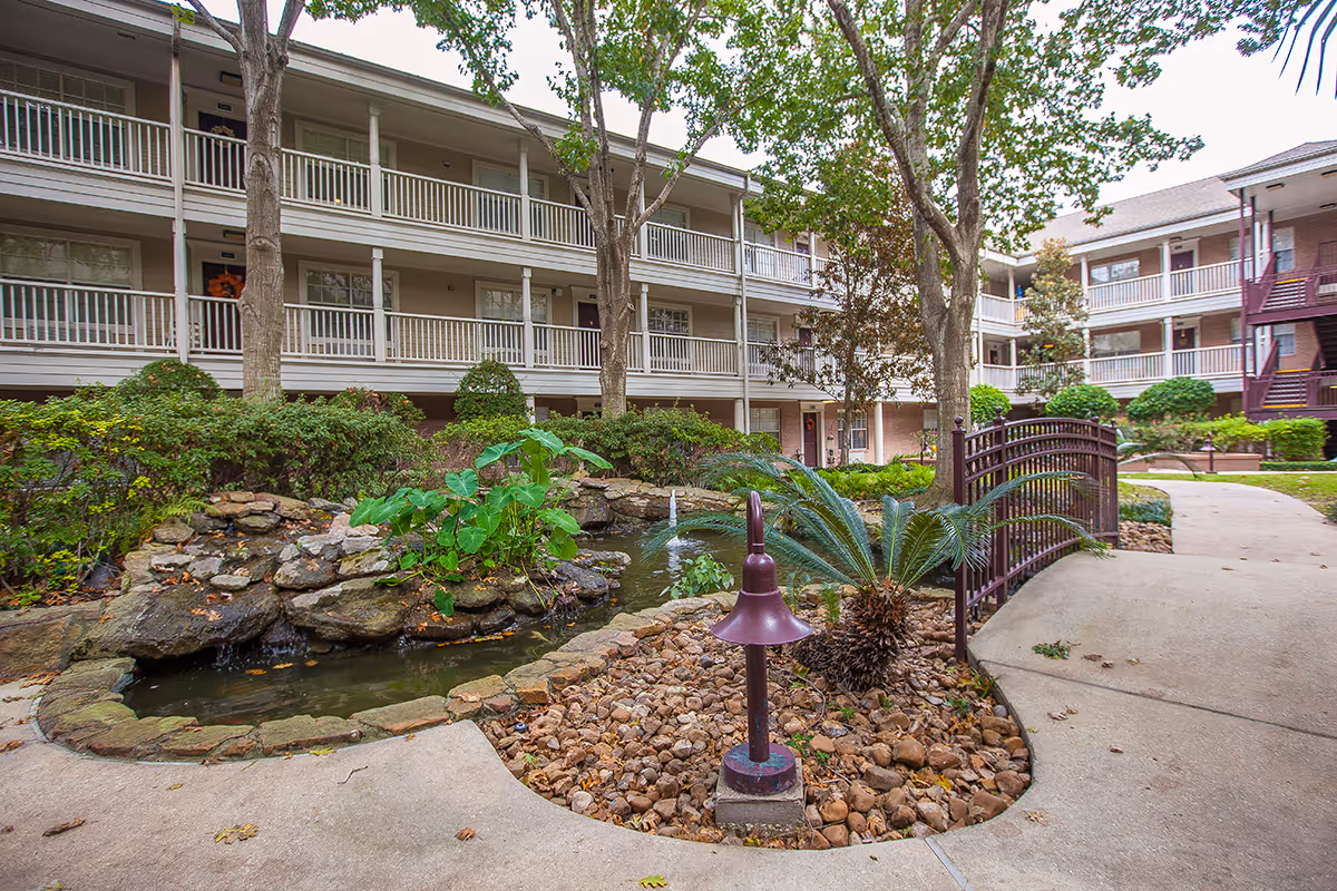 Courtyard of a multi-story senior living building with balconies, a small pond and waterfall, a footbridge and landscaped walkways.