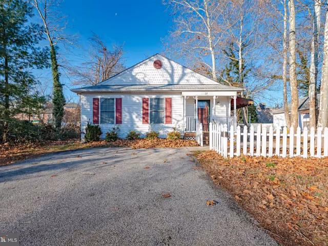 A small white house with red shutters and a red door, surrounded by leafless trees and a white picket fence on the right side. The driveway is paved and there are dry leaves scattered on the ground. The sky is clear and blue.