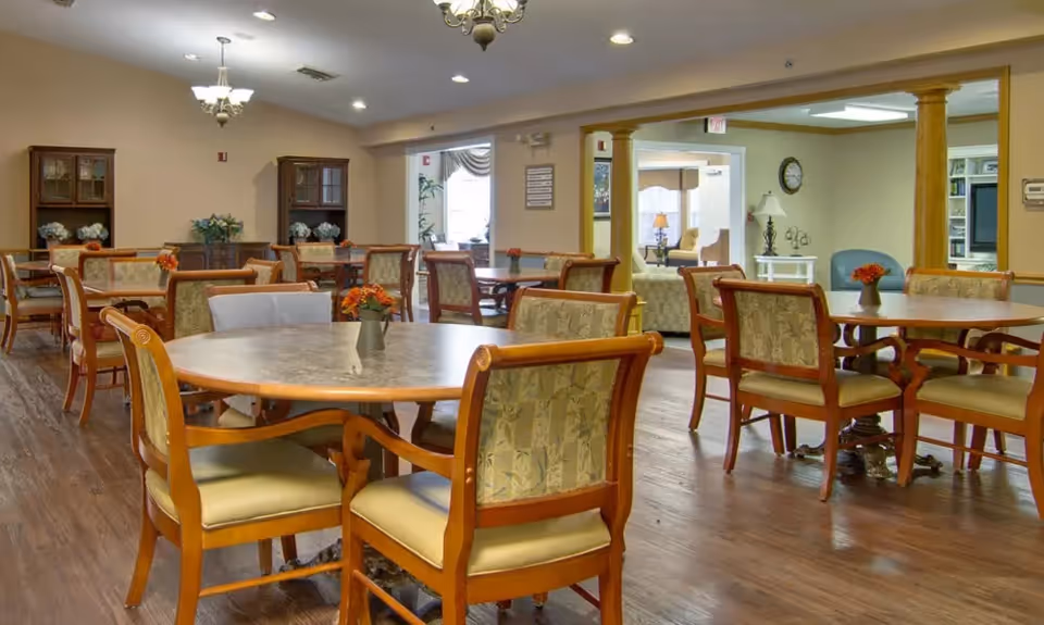 Spacious dining room with round tables, wooden chairs, floral centerpieces, and wood floors in a senior living facility.