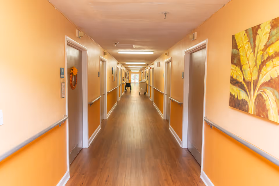 A long, well-lit hallway in a senior living facility with orange walls and wooden flooring. Doors line both sides of the corridor, each with handrails along the walls. A painting with yellow leaves is visible on the right wall, and a wreath hangs on one of the doors on the left. At the end of the hallway, there is a window letting in natural light.