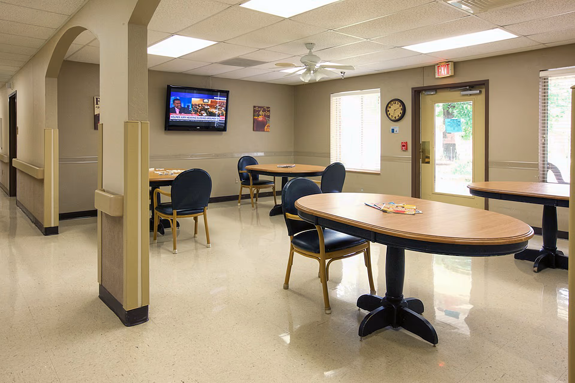 A well-lit common area with several wooden tables and blue cushioned chairs arranged around them. A flat-screen TV is mounted on the beige wall, showing a news broadcast. There are windows letting in natural light, a clock on the wall, and an exit door leading outside. The floor is shiny and light-colored, and the ceiling has recessed lighting and a ceiling fan.