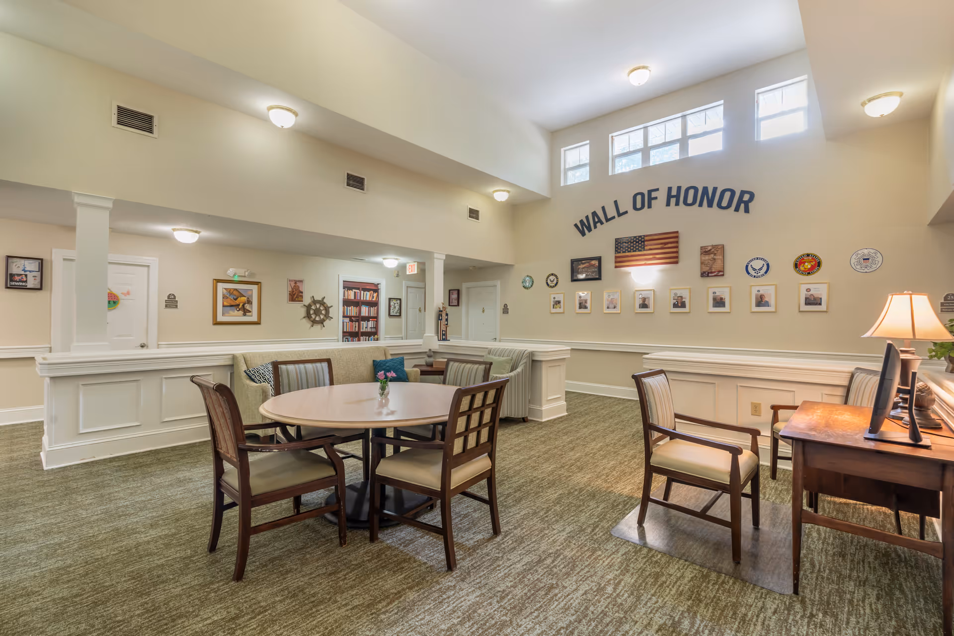 A well-lit common area in a senior living facility featuring a round table with four chairs, a small desk with a computer and two chairs, and a wall labeled 'Wall of Honor' displaying an American flag and framed photos. The room has beige walls, carpeted floor, and several light fixtures on the ceiling.