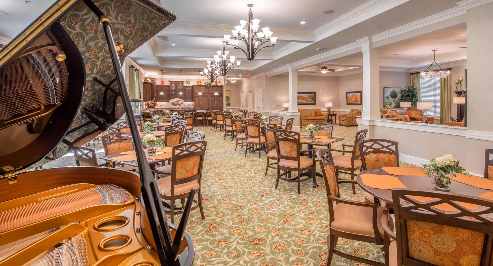 Interior dining and common area with multiple round tables and chairs, a grand piano in the foreground, and lounge seating in the background.