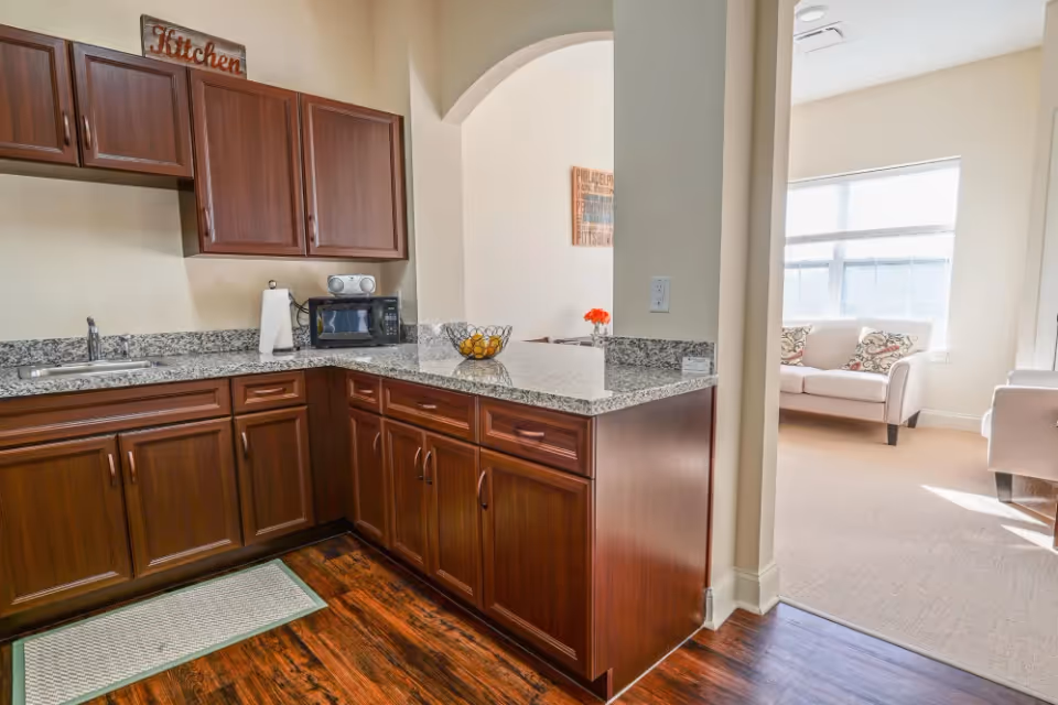 A bright kitchen area with dark wooden cabinets and granite countertops. There is a sink, a microwave, a paper towel holder, and a decorative bowl with fruit on the counter. Above the cabinets, a sign reads 'Kitchen'. The kitchen opens into a living room area with beige carpeting, a large window letting in natural light, and white upholstered chairs with patterned cushions.