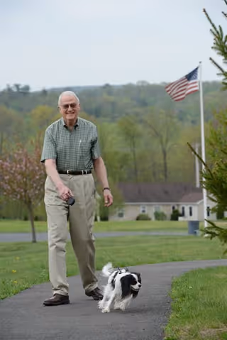 An elderly man wearing a green checkered shirt and beige pants is walking a small black and white dog on a leash along a paved path in a grassy outdoor area. In the background, there is a building, trees, and an American flag on a flagpole.