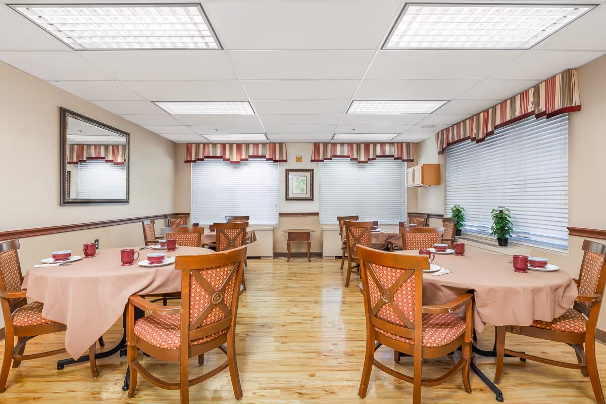 A dining room with several round tables covered with light brown tablecloths. Each table is surrounded by wooden chairs with patterned upholstery. The tables are set with red cups and white plates. The room has large windows with white blinds and striped valances. There is a large mirror on one wall and a framed picture between two windows. The floor is wooden, and the ceiling has fluorescent lighting panels.