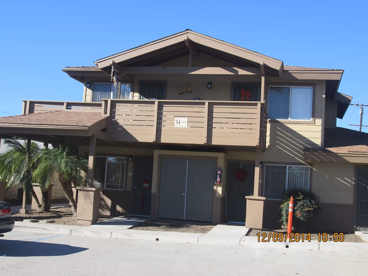 Exterior view of a two-story residential building with beige siding and brown trim. The building has a covered entrance, balcony with railing, several windows, and a few small palm trees near the entrance. The sky is clear and blue.