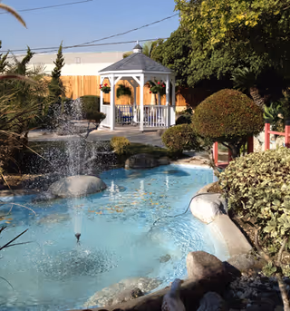 A peaceful outdoor garden area featuring a small pond with a water fountain, surrounded by trimmed bushes and trees. In the background, there is a white gazebo with hanging flower baskets and chairs inside. The area is enclosed by a wooden fence under a clear blue sky.