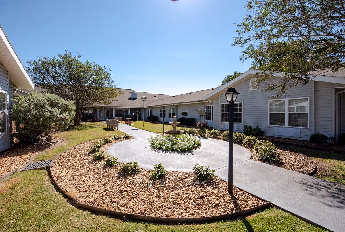 Outdoor courtyard area at Trustwell Living at Bailey Place featuring a circular flower bed with a small fountain in the center, surrounded by a paved walkway, lamp posts, trees, bushes, and single-story building structures under a clear blue sky.