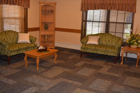 A cozy living room with two green patterned sofas, each with a white pillow. There is a wooden coffee table in the center with a decorative bowl on it. A wooden corner shelf with the word 'LAUGH' on the top shelf stands against the wall. The room has large windows with brown curtains and blinds, and a small wooden side table with a floral arrangement is placed next to one sofa. The floor is carpeted with a patterned design.