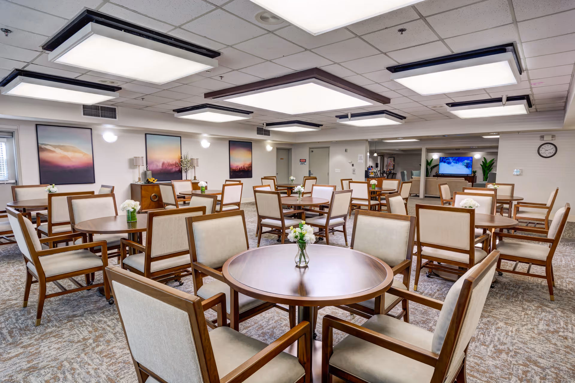A spacious senior living dining room with multiple round wooden tables surrounded by beige cushioned chairs. Each table has a small vase with white flowers. The room is well-lit with large rectangular ceiling lights and has framed landscape artwork on the walls. A television and some plants are visible in the background.
