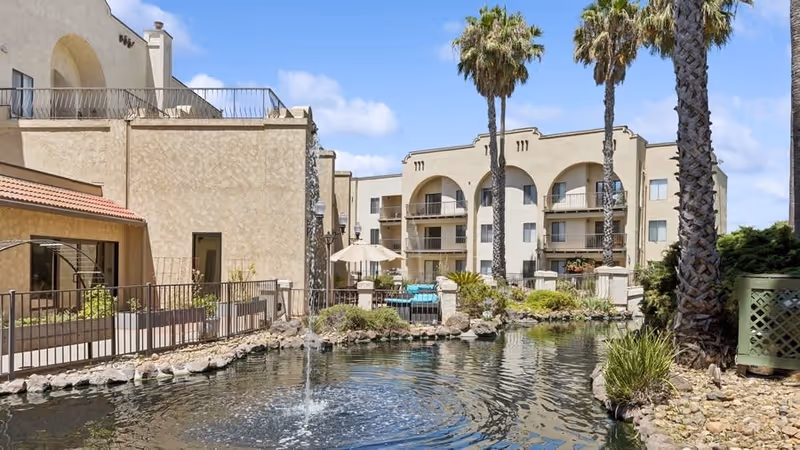 Outdoor view of a senior living facility with beige multi-story buildings, balconies, tall palm trees, a pond with a small fountain, and landscaped greenery under a partly cloudy sky.