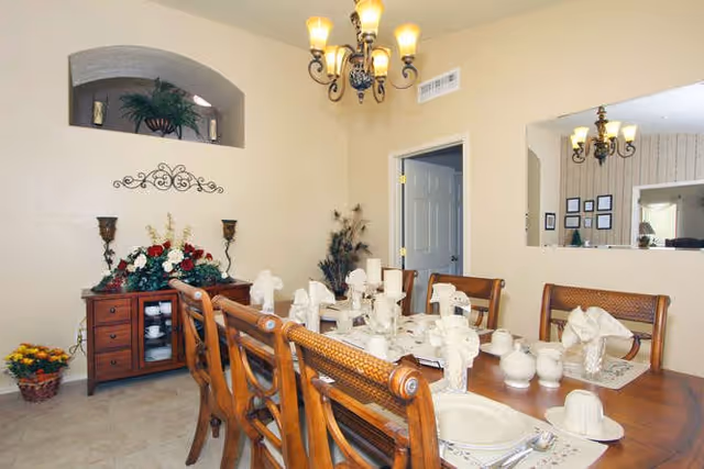 A dining room with a wooden dining table set for eight people, featuring white napkins folded in a decorative manner, white plates, cups, and silverware. The room has beige walls, a chandelier hanging from the ceiling, a wooden sideboard with floral decorations, and a small window-like opening in the wall showing another chandelier in an adjacent room.