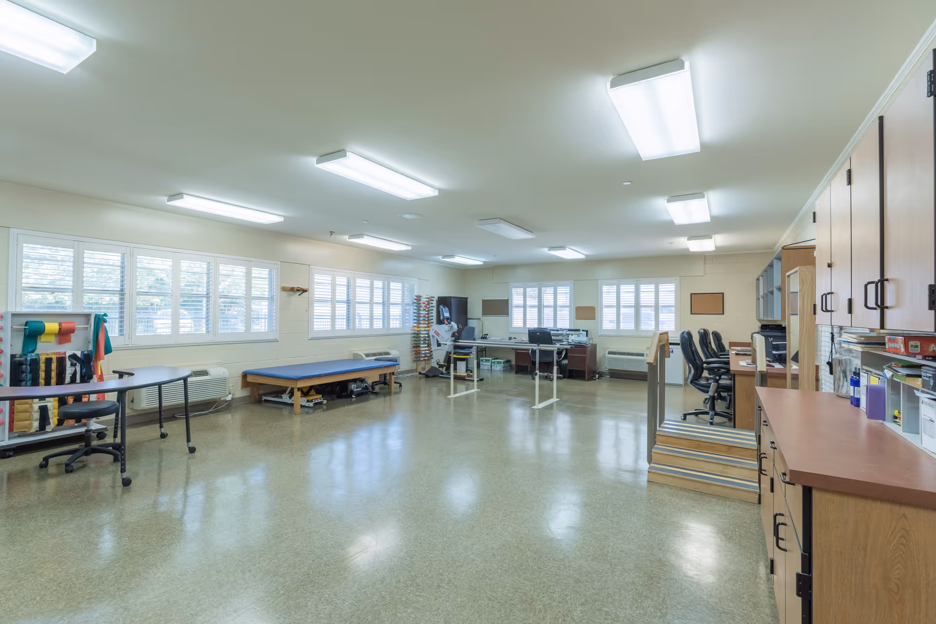 A large, bright interior therapy/activity room with treatment tables, exercise equipment, desks, cabinets, and chairs.
