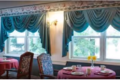 A dining area with tables covered in pink tablecloths, set with plates, cups, and a vase with yellow flowers. The room has large windows with teal draped curtains and floral wallpaper border near the ceiling.