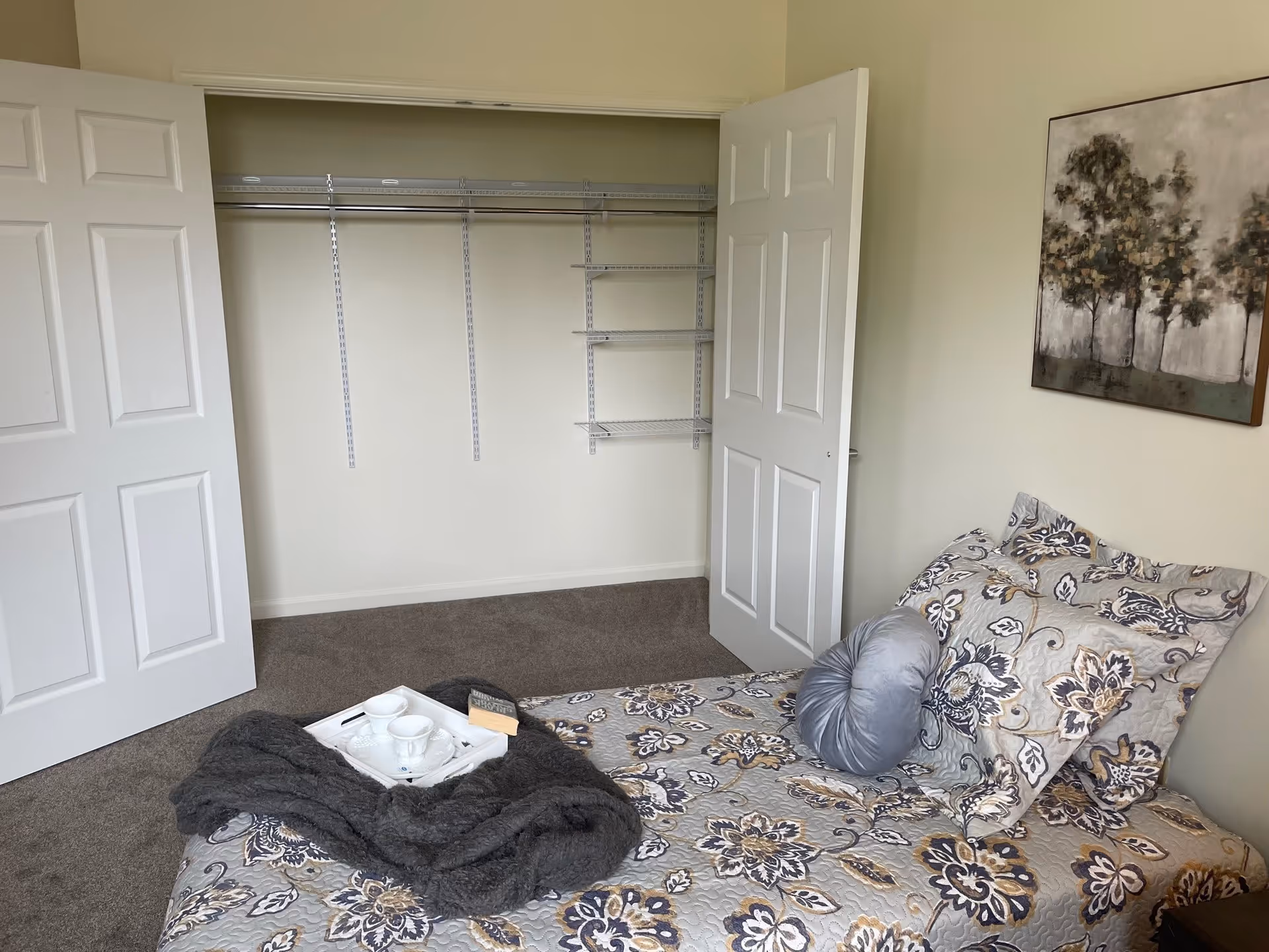 A bedroom with a bed covered in a floral patterned quilt and multiple pillows, including a round gray pillow. A dark gray blanket and a white tray with two teacups and a book are on the bed. The room has an open closet with white double doors and built-in shelving. A painting of trees hangs on the wall above the bed.