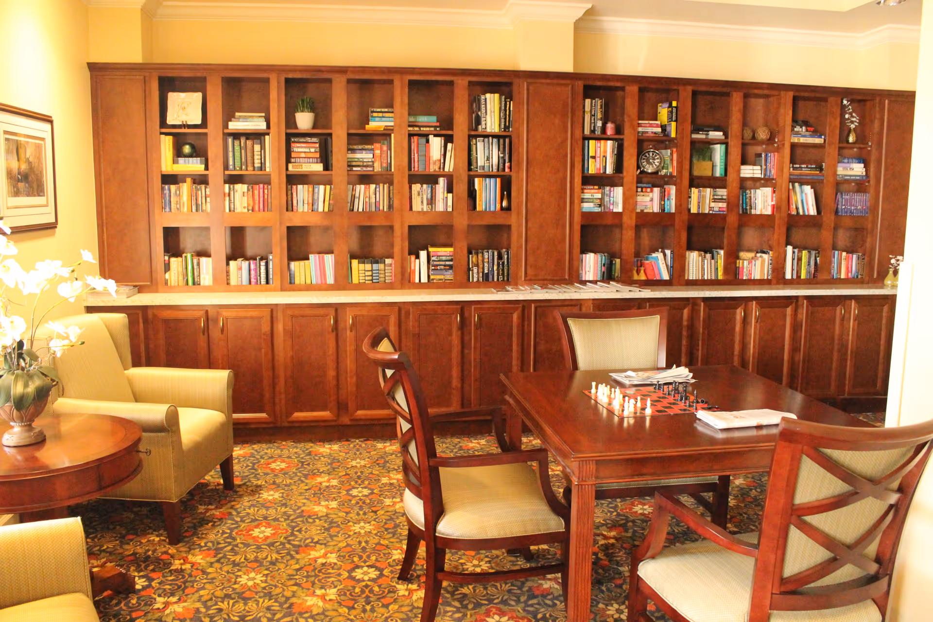 A cozy reading and game room with wooden bookshelves filled with books along the back wall, a wooden table with chairs around it, and a chessboard set up on the table. There are comfortable armchairs and a small round table with a potted plant on it. The room has warm lighting and a patterned carpet.