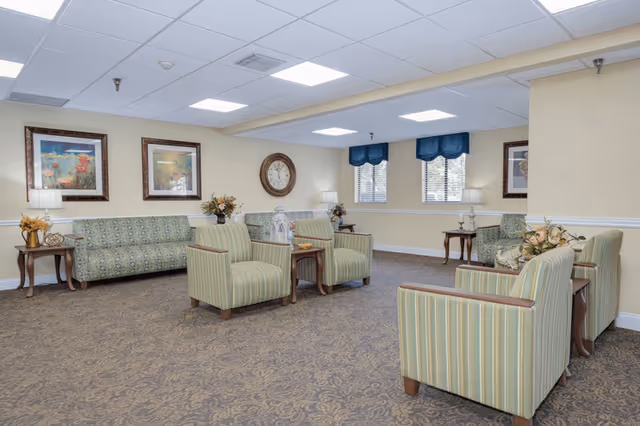 Spacious senior living common room with upholstered armchairs and sofas arranged around side tables, wall art, and windows.