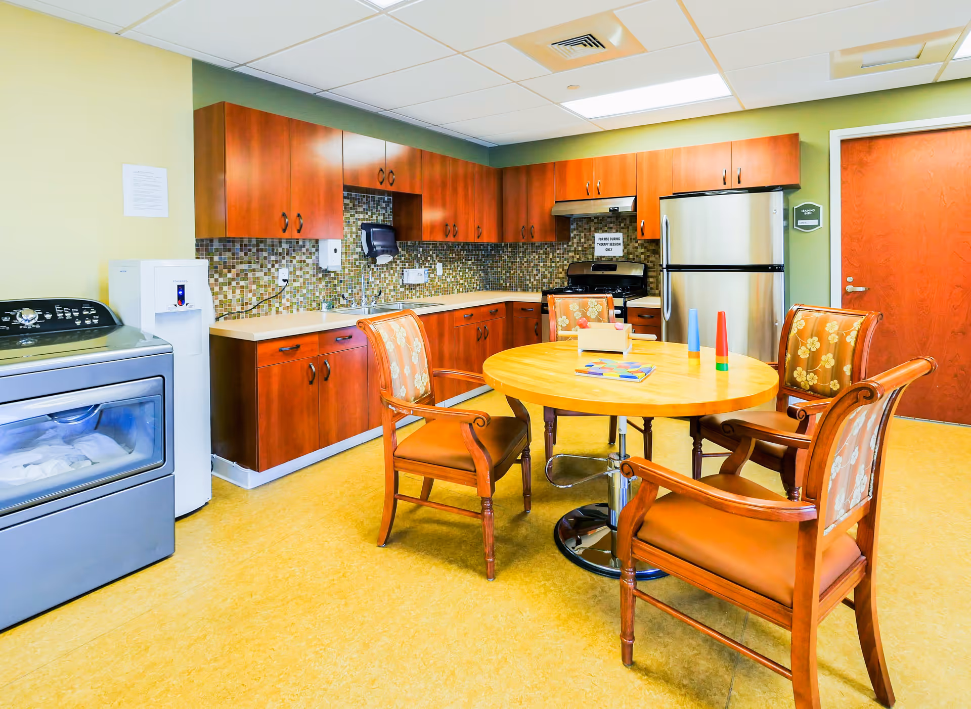 A bright kitchen area with wooden cabinets, a mosaic tile backsplash, a stainless steel refrigerator, stove, and a washing machine. In the center, there is a round wooden table with four wooden chairs that have floral upholstery. The floor is yellow, and the walls are painted green and beige. There are some colorful objects on the table and a closed wooden door in the background.