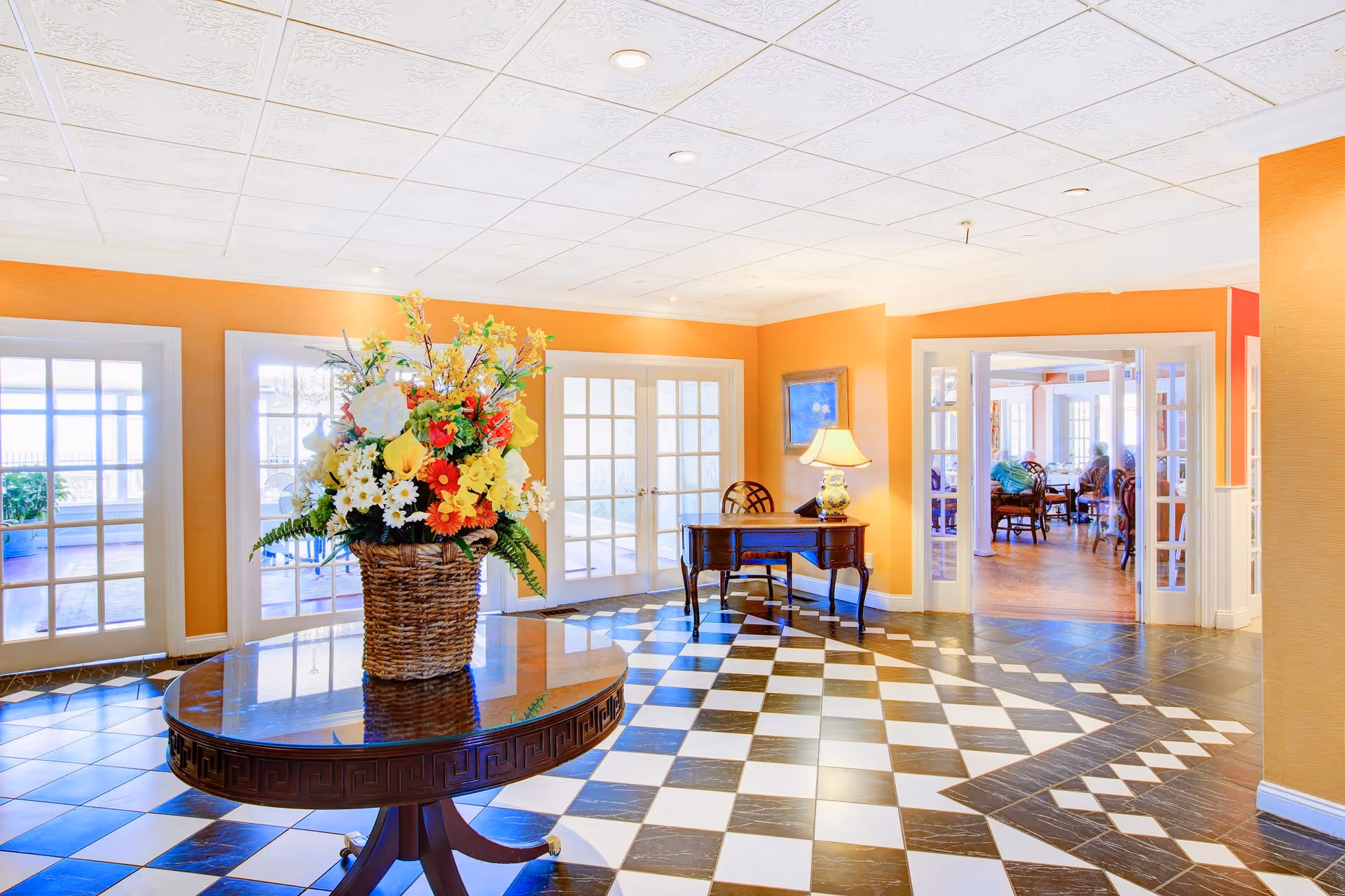 Bright and spacious interior area with black and white checkered tile flooring, a round wooden table with a large basket of colorful flowers, orange walls, and multiple glass-paneled doors leading to other rooms. A wooden desk with a lamp and chair is positioned against one wall, and through an open doorway, a sitting area with chairs and people is visible.