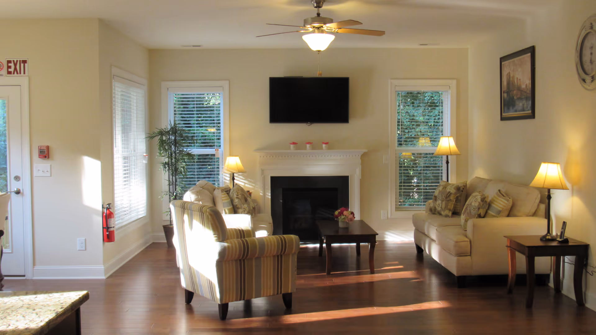 Well-lit living room with sofas and armchairs arranged around a fireplace and wall-mounted TV, with lamps and hardwood floors.