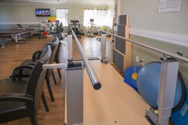 A rehabilitation or physical therapy room with parallel bars for walking exercises, several black chairs lined up along the left side, exercise equipment including a large blue exercise ball and balance pads on the right, and a treadmill and stationary bike near a window in the background.