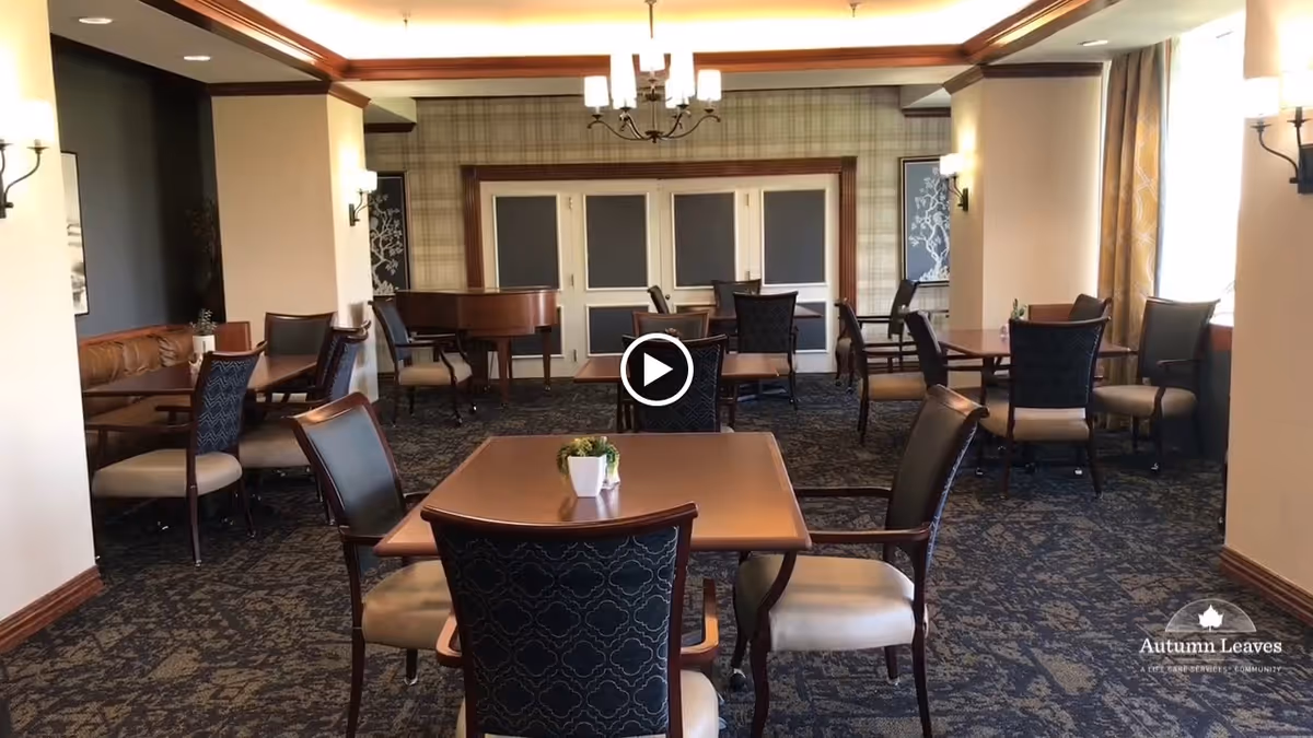 A well-lit dining room with multiple tables and chairs arranged neatly. The room features patterned carpet, a chandelier, wall sconces, and large windows with curtains allowing natural light. A piano is visible in the back corner, and there are framed artworks on the walls.
