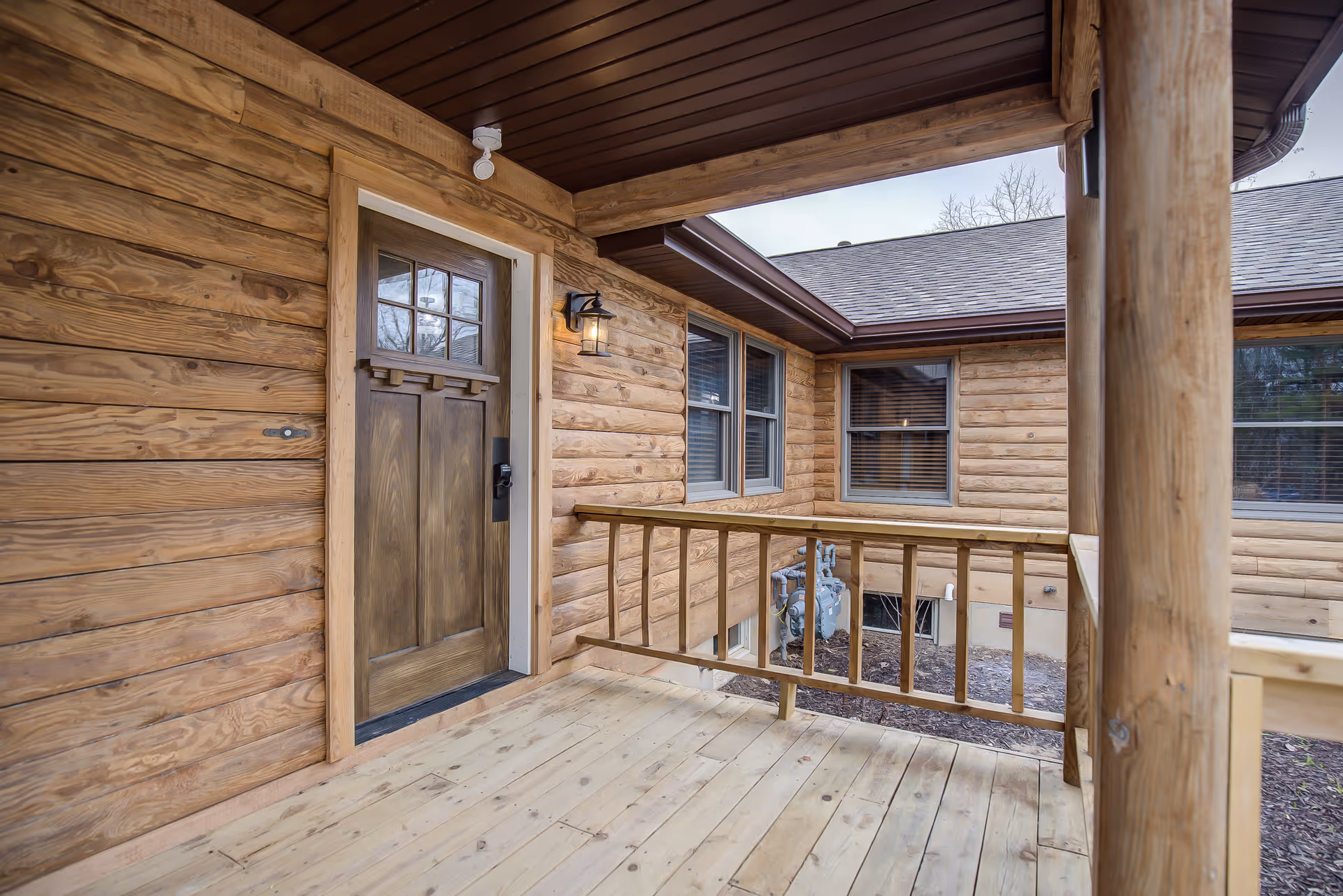 Covered wooden porch with a rustic wooden entry door, railing, and adjacent log-style exterior walls and windows.