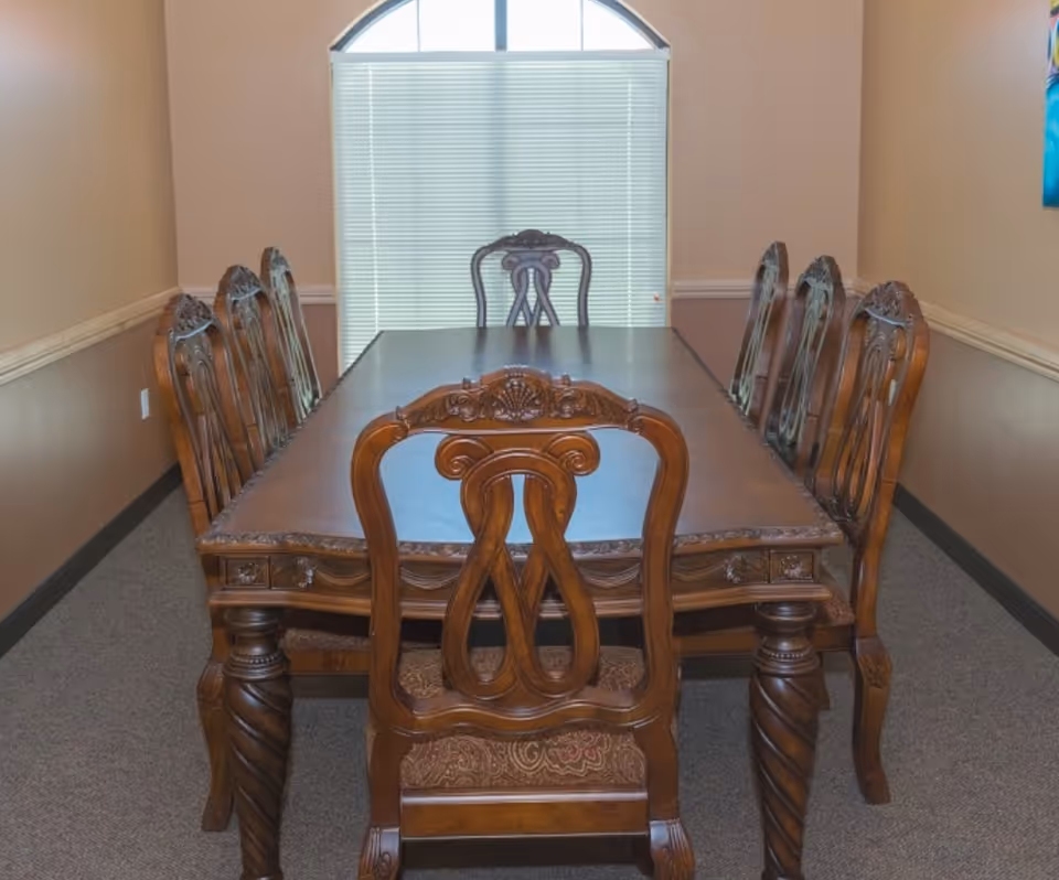 A formal dining room with a large rectangular wooden table surrounded by eight ornate wooden chairs. The room has beige walls, a carpeted floor, and a large window with closed blinds and an arched top letting in natural light.