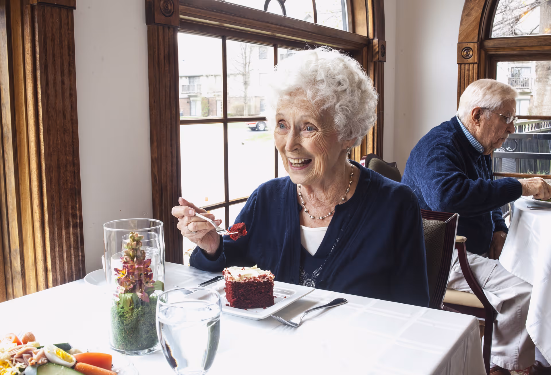 An elderly woman smiling at a dining table eating a slice of cake with another older man seated behind her.