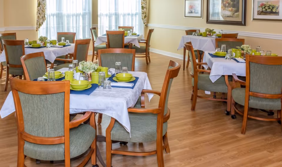 A dining room with several tables covered in white tablecloths and set with green plates, bowls, cups, and clear drinking glasses. Each table has wooden chairs with green upholstery, and there are floral centerpieces on the tables. The room has light-colored walls with framed artwork and large windows with sheer curtains allowing natural light to enter.